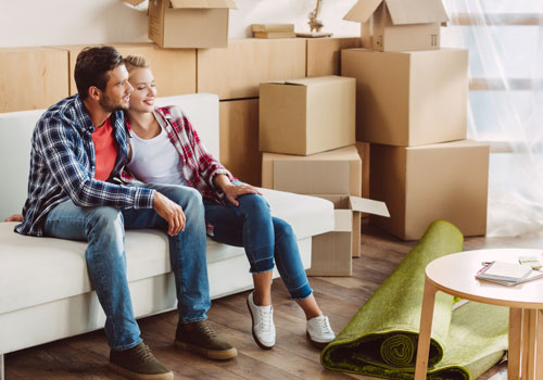 A man and a woman sitting on a couch with several moving boxes behind them